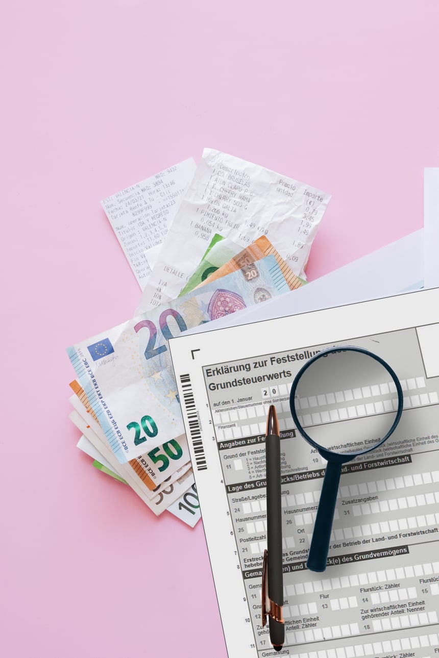 Bild von einem Formular zur Grundsteuererklärung. mit einem pinken Hintergrund und ringsrum Geldscheine in Euro mit einem Kugelschreiber sowie einer Lupe.